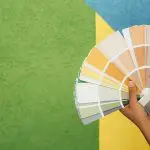 Hands holding a fan of paint color swatches in front of a colorful painted wall, illustrating how to choose interior paint colors for your home.