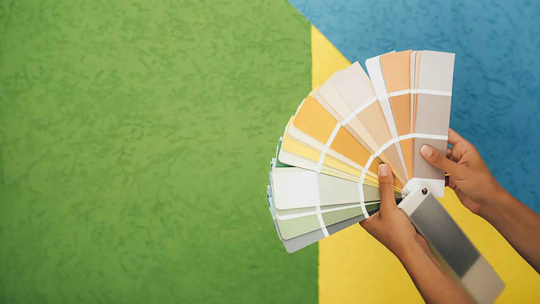 Hands holding a fan of paint color swatches in front of a colorful painted wall, illustrating how to choose interior paint colors for your home.