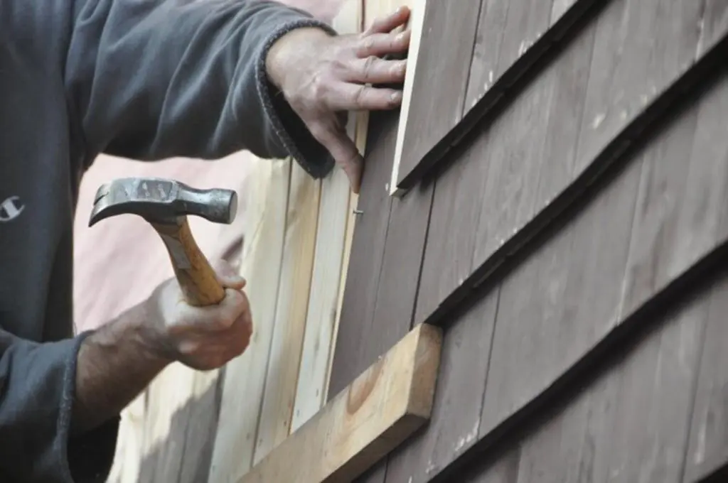 A carpenter hammers a nail into fresh trim while replacing damaged cedar shingle siding, one of the common carpentry repairs needed before painting.
