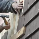 A carpenter hammers a nail into fresh trim while replacing damaged cedar shingle siding, one of the common carpentry repairs needed before painting.