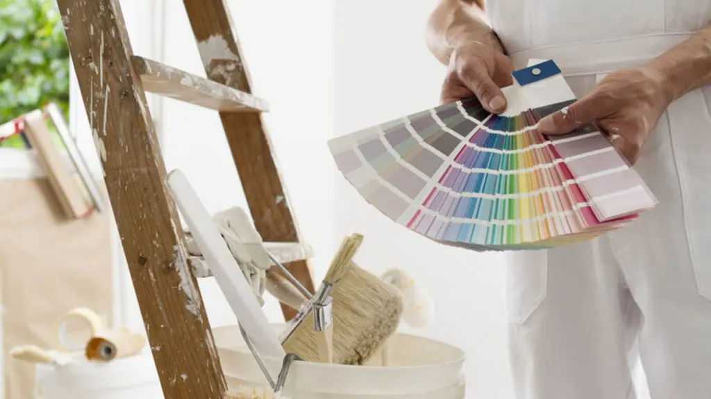A painter holds a fan of paint color swatches next to a ladder and supplies, one of the key decisions homeowners should sort out before interior painting begins.