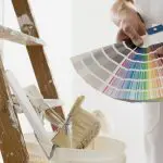 A painter holds a fan of paint color swatches next to a ladder and supplies, one of the key decisions homeowners should sort out before interior painting begins.